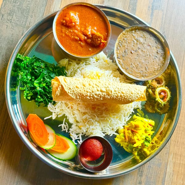 Thali platter with basmati rice, papad, rich curry, dal, saag, bitter gourd, salad, and gulab jamun on a steel plate.