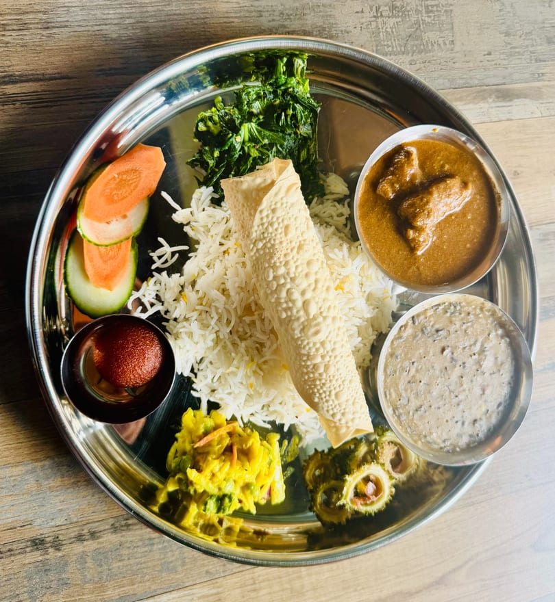 Nepalese thali platter with basmati rice, papad, curry, dal, saag, cauliflower, pickles, and gulab jamun on a steel plate.
