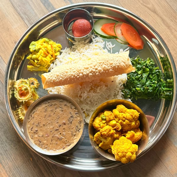 Thali platter with rice, papad, curried cauliflower, dal, saag, mixed vegetables, and gulab jamun on a steel plate.